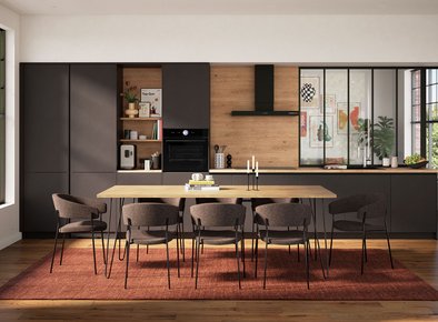 Cuisine moderne élégante avec des armoires noires mates et bois, une grande table en bois entourée de chaises rembourrées, et un éclairage naturel mettant en valeur une verrière et une crédence chaleureuse.